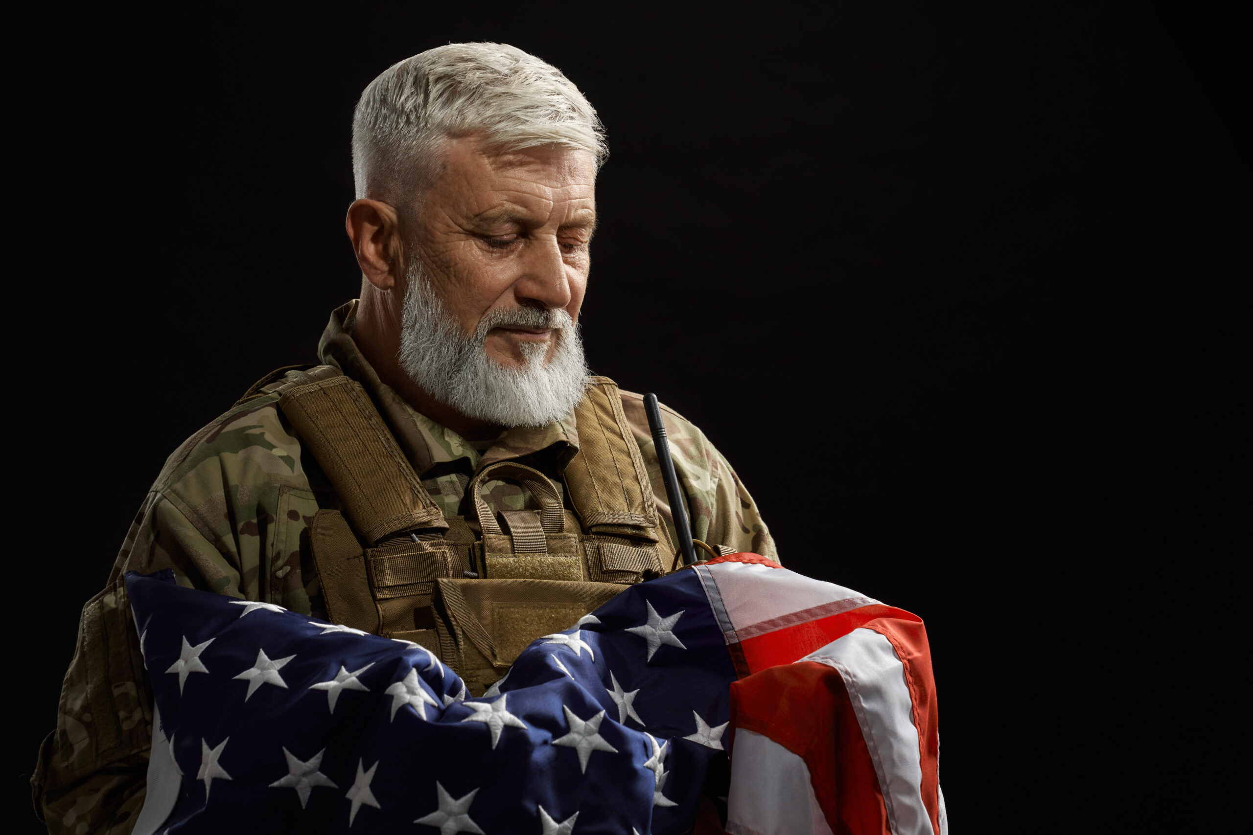 Front view of bearded american military veteran holding flag in arms. Portrait of old proud male officer in camouflage uniform posing in dark atmosphere. Concept of military, patriotism.