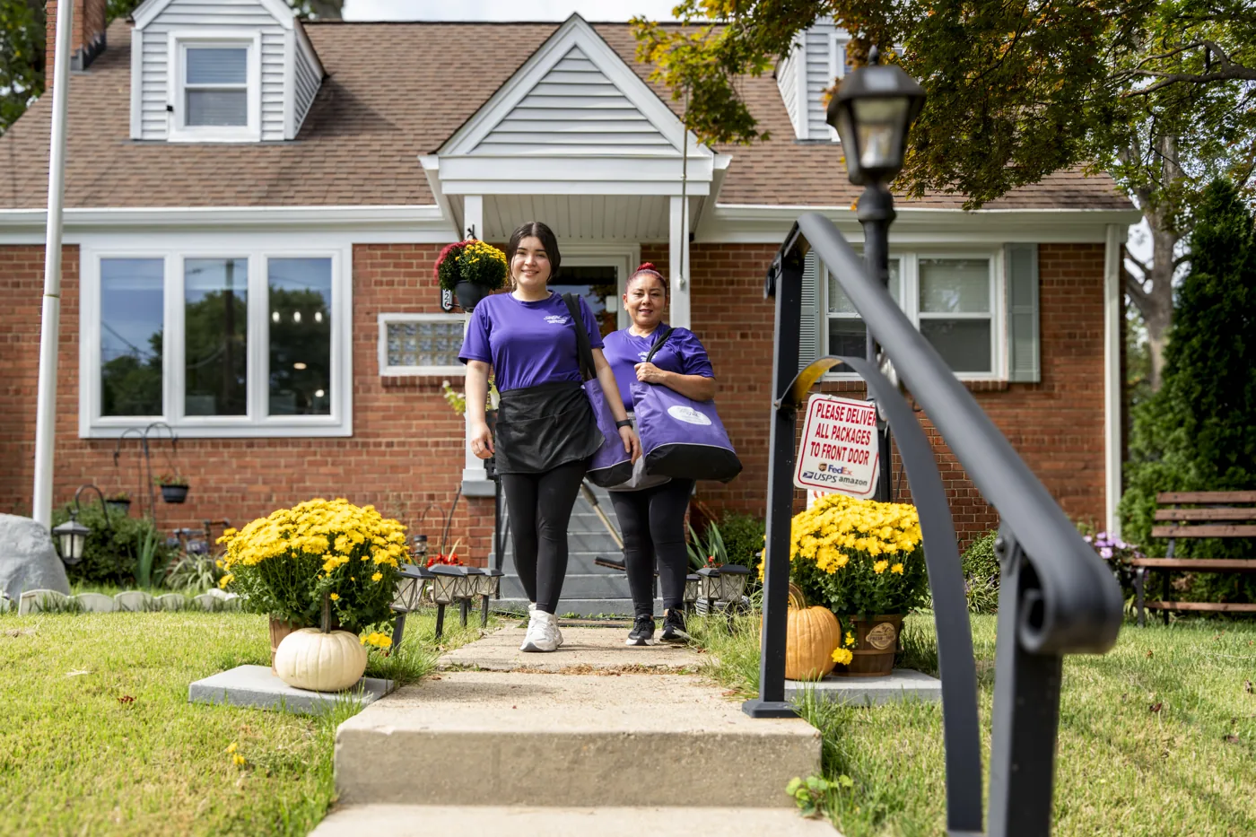 Two All Neat cleaning professionals carrying supplies outside a brick home